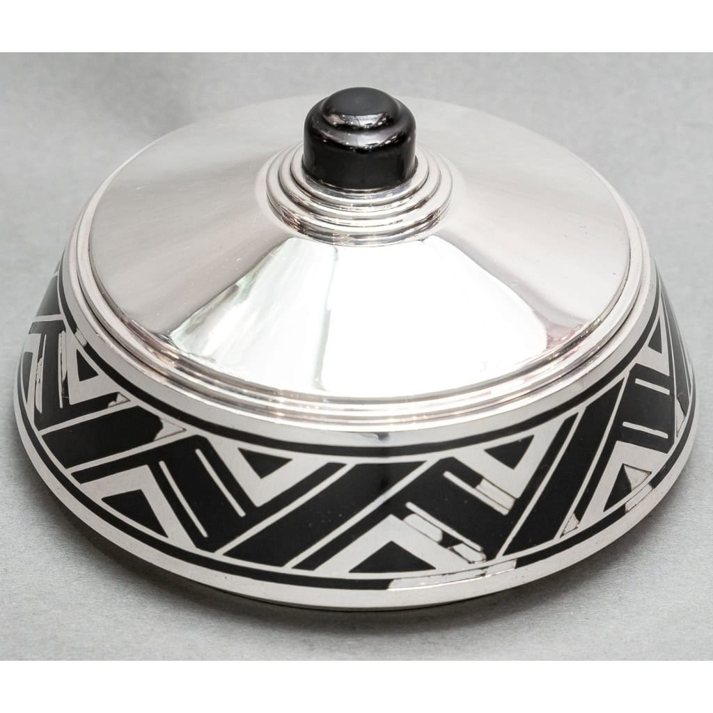 Orfevre R.Linzeler – Boite en argent massif et émail noir circa 1930 | Biron Orfèvre R. LINZELER – Boite en argent massif et émail noir – Epoque ART DECO Rare boite ronde couverte en argent et émail noir dans un décor stylisé ART DECO. L’intérieur est en vermeil et le couvercle est surmonté d’une prise en ébène. (Petits manques à l’émail). Dimensions : diamètre 14 cm – hauteur 8 cm env. Matière : Argent 1er titre 950/°°° Poids : 345 grammes Poinçon : MINERVE Epoque : ART DECO vers 1930 Orfèvre : Robert LINZELER Origine : France/Paris