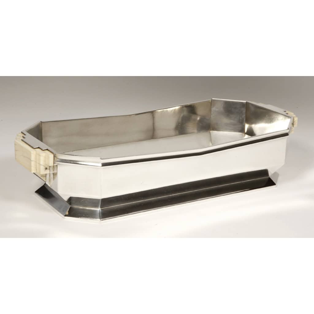 Orfèvre TETARD – Jardinière en argent massif époque ART DECO 1930 | Biron Orfèvre TETARD - Jardinière en argent massif époque ART DECO 1930 Jardinière en argent massif de forme octogonale découpe latérale en V, montée sur une bâte à piédouche, cet objet est flanqué de 2 anses à décor de gradins, rivetées sur la jardinière elle-même. Objet de maitrise dessiné par Valéry BIZUARD (1875-1945), designer de la Maison Tétard Orfèvre : TETARD Frères Cette JARDINIERE est répertoriée dans la revue ; "MOBILIER ET DECORATION 1930" Revue française des Arts Décoratifs Appliqués Dimensions : long 34,2 cm - largeur : 15,8 cm - hauteur 7,3 cm Poids : 1040 grammes Matière : Argent 1er titre 950/°°° Poinçon : Minerve Orfèvre : TETARD à PARIS Origine : France/Paris