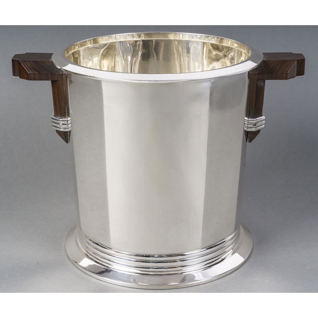 PUIFORCAT – Seau à Champagne « Bayonne » en argent massif ART DECO | Biron PUIFORCAT – Seau à Champagne "Bayonne"en argent massif ART DECO Important Seau à Champagne en argent massif, à décor de deux sections de pans coupés, séparées par des arrondis sur lesquels sont fixées les anses en bois de forme géométrique et renforcées par des joncs filetés en argent. au modèle Bayonne . Le corps uni est cintré d’un bandeau cerclé des trois joncs à la base. Dimensions : Hauteur 21,5 cm – Largeur au col 21,5 cm Largeur aux anses 29,5 cm Matière : Argent 1er titre 950/°°° Poids : 2 715 grammes Poinçon : MIINERVE Epoque : Puiforcat Paris – Edition 1985 d’après un modèle créé en 1935 par Jean Elysée Puiforcat (1897-1945) Orfèvre : JEAN E. PUIFORCAT à PARIS Origine : France/Paris JEAN PUIFORCAT par Françoise de Bonneville page 82 (saucière au modèle)