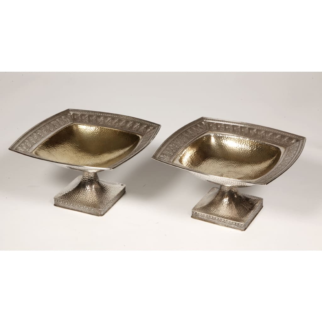 GORHAM – Paire de Coupes en argent massif martelé Birmingham XXe | Biron GORHAM – Paire de Coupes en argent massif martelé Birmingham XXe Très belle paire de coupes en argent martelé montée sur une base carrée à décor de festons surmontée d’un fût supportant une coupe mi creuse vermeillée, bordée d’une ciselure d’ogives fleuries ponctuées de gerbes de blé, le tout dans une forme carrée légèrement arrondie. Dimensions : hauteur 13,5 cm – base 11 cm x11 cm – coupe 24x24 cm Matière : Argent 925/°°° Poids : 1486 grammes Poinçons : Anglais lion passant – ancre Birmingham – N° ordre 1722 Epoque : début XXe Orfèvre : C. CODMAN pour GORHAM Origine : BIRMINGHAM En 1891, l’anglais William C. CODMAN s’est vu confier la responsabilité du département Projets, il créa en 1897 la série « MARTELE ATHENIC » pour la grosse orfèvrerie. En s’inspirant des modèles antiques similaires à nos coupes. La gamme comportait des vases, des coupes, des timbales et des services à thé. La production s’est poursuivie jusqu’en 1910