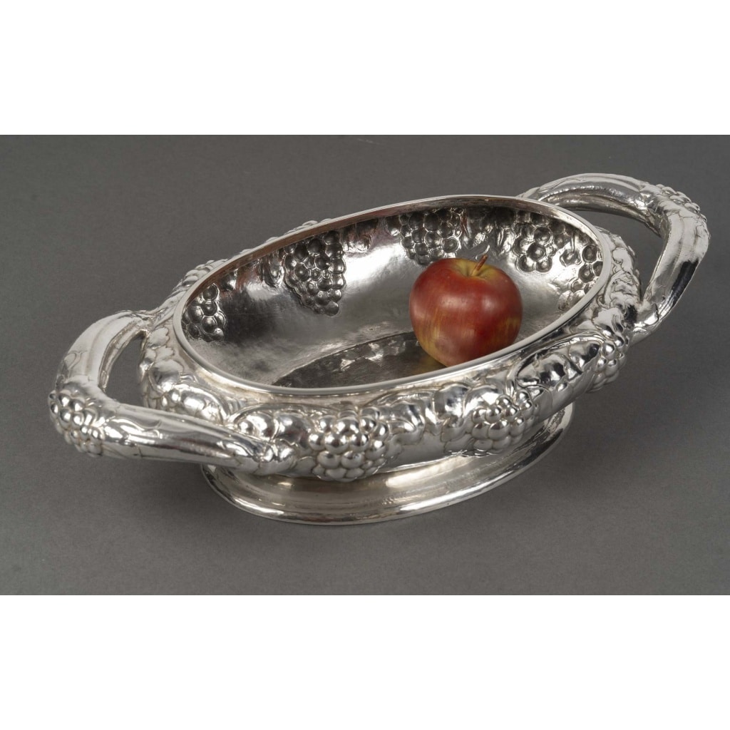 JEAN SERRIERE – Jardinière ovale en argent massif XXe vers 1920 | Biron JEAN SERRIERE – Jardinière ovale en argent massif XXe vers 1920 JARDINIERE ovale en argent massif dite « Aux raisins » posant sur un piédouche à légère doucine à décor de pampres repris sur les anses. Sous la base poinçon au vase étrusque de « A. HEBRARD/PARIS », Dimensions : longueur 49 cm – largeur 25 cm Matière : Argent 1er titre 950/°°° Poids : 2.060 grammes Poinçon ;Tête de sanglier Epoque : XXe vers 1920 Orfèvre : JEAN SERRIERE (1893 – 1968) Signature et Poinçon d’A.HEBRARD, Paris Provenance : Ancienne collection LANVIN Origine : France/Paris Bibliographie : Jean Serrière (1893-1968) est né à Nancy le 26 février 1893, dans une famille lorraine. Son père, Émile Serrière, était graveur, et sa mère, Adèle Serrière, a dirigé la fonderie Hébrard après le décès de René Desbois en 1909. En 1900, Jean Serrière s’installe à Paris pour ses études et fréquente des ateliers prestigieux, notamment celui de Georges Desvallières. Après la Première Guerre mondiale, il se tourne vers la dinanderie, travaillant le cuivre et l’argent martelé, et crée des meubles qu’il expose au Pavillon de Marsan. Autodidacte et curieux, il s’intéresse ensuite à la technique des émaux, ajoutant ainsi le métier d’émailleur à ses compétences artistiques. Il reçoit divers prix et distinctions lors des expositions de 1925 et 1937, côtoyant les plus grands artistes de son temps. La fonderie Hébrard, fondée par Adrien-Aurélien Hébrard en 1902, était réputée pour la qualité de ses bronzes réalisés selon la technique de la cire perdue. Après la mort de René Desbois en 1909, Adèle Serrière, la mère de Jean Serrière, a pris la direction de la fonderie. En 1925, Adrien Hébrard confie la fonderie à sa fille Nelly, qui la dirige officiellement à partir de 1932 jusqu’à sa liquidation en 1934. Dans les années 1920, Jean Serrière collabore avec la fonderie Hébrard pour la réalisation de ses œuvres en dinanderie et en émail. Par exemple, une aiguière en argent créée par Jean Serrière et fondue par Hébrard vers 1930 témoigne de cette collaboration.