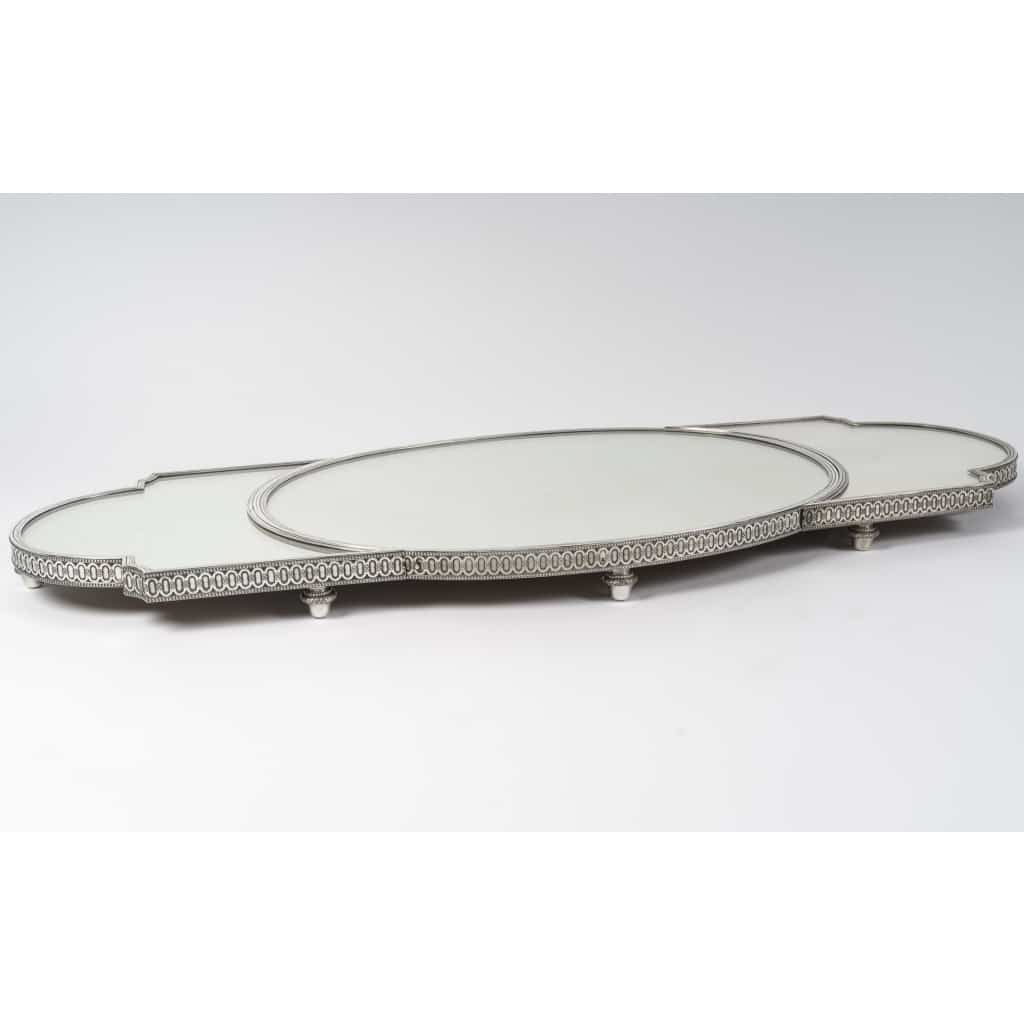 ODIOT Surtout de table en argent 3 parties, Napoléon III, fin XIXe | Biron ODIOT – Surtout de table en argent 3 parties, Napoléon III, fin XIXe Important surtout de table en trois parties, à fond de miroirs sur un parquet de chêne, cernées d’une lingotière en argent massif à décor d’oves et rang de fines perles, montées sur des pieds toupies. Le centre ovale est complété par deux parties qui s’encastrent ou s’écartent selon l’usage. Dimensions : Partie centrale : 61 x 44,5 cm Longueur maxi : 107,5 cm x 44,5 cm Hauteur : 6 cm Matières : Argent 1er titre 950/1°°°, chêne, miroirs Poinçon : MINERVE Epoque : FIN XIXe Orfèvre : ODIOT/ Prévost & Cie (1895-1905) Origine : France/Paris