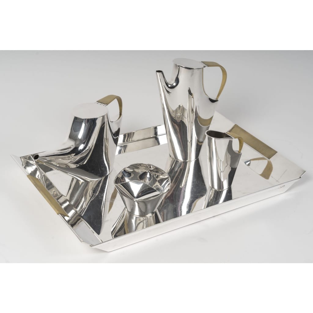 CHRISTOFLE – SABATTINI – Set Thé/Café Moderniste sur son plateau Argent Massif Circa 1957/1968 | Biron CHRISTOFLE-CARDEILHAC & Lino SABATTINI (né en 1925), designer. Modèle COMO, créé vers 1956-1957 Service à thé et café moderniste en argent massif comprenant 5 pièces : cafetière, théière, sucrier et pot à lait en argent uni et un Plateau rectangulaire aux anses gainées de fibres à l'imitation du raphia. Dimensions : Théière : Longueur : 30 cm Base : 7 x 10 cm Hauteur : 13,5 cm Cafetière : Longueur : 16 cm - Base : 6,5 cm - Hauteur : 21,5 cm Crémier : Longueur :9 cm- Base : 4,5 cm x 3,5 cm - Hauteur : 12 cm sucrier : 10 x 10 cm Base : 5,5 cm Hauteur : 7 cm Plateau : 49 x 30 cm Hauteur : 7 cm Matière : Argent 1er titre 940/1°°° Poids : 3 Kg 606 Poinçon : ARGENTO 940/1000 Orfevre : CHRISTOFLE Origine : Au milieu des années 1950, Lino Sabattini ouvre un atelier d'orfèvrerie à Milan qui attire rapidement l'attention de Gio Ponti. Ce dernier lui consacre un article dans le magazine Domus et présente ses oeuvres en 1956 à Paris dans une exposition consacrée au design italien. Sabattini obtient dès lors une reconnaissance internationale et devient directeur du design pour Christofle Orfèvrerie (qui a racheté Cardeilhac en 1951). Il insuffle un vent de nouveauté dans cette maison fondée en 1830, en imaginant des couverts, pièces de forme et bougeoirs aux lignes modernistes pour la collection " Formes nouvelles". Le modèle Como est le plus souvent réalisé en métal argenté par Christofle pour la collection Gallia. Cette édition en argent par Christofle-Cardeilhac est donc particulièrement rare. Provenance : Acquis directement auprès de la maison Christofle le 12 juin 1957. Bibliographie : Connaissance des Arts, mai 1999, n°561 p. 71 pour un modèle identique en argent.
