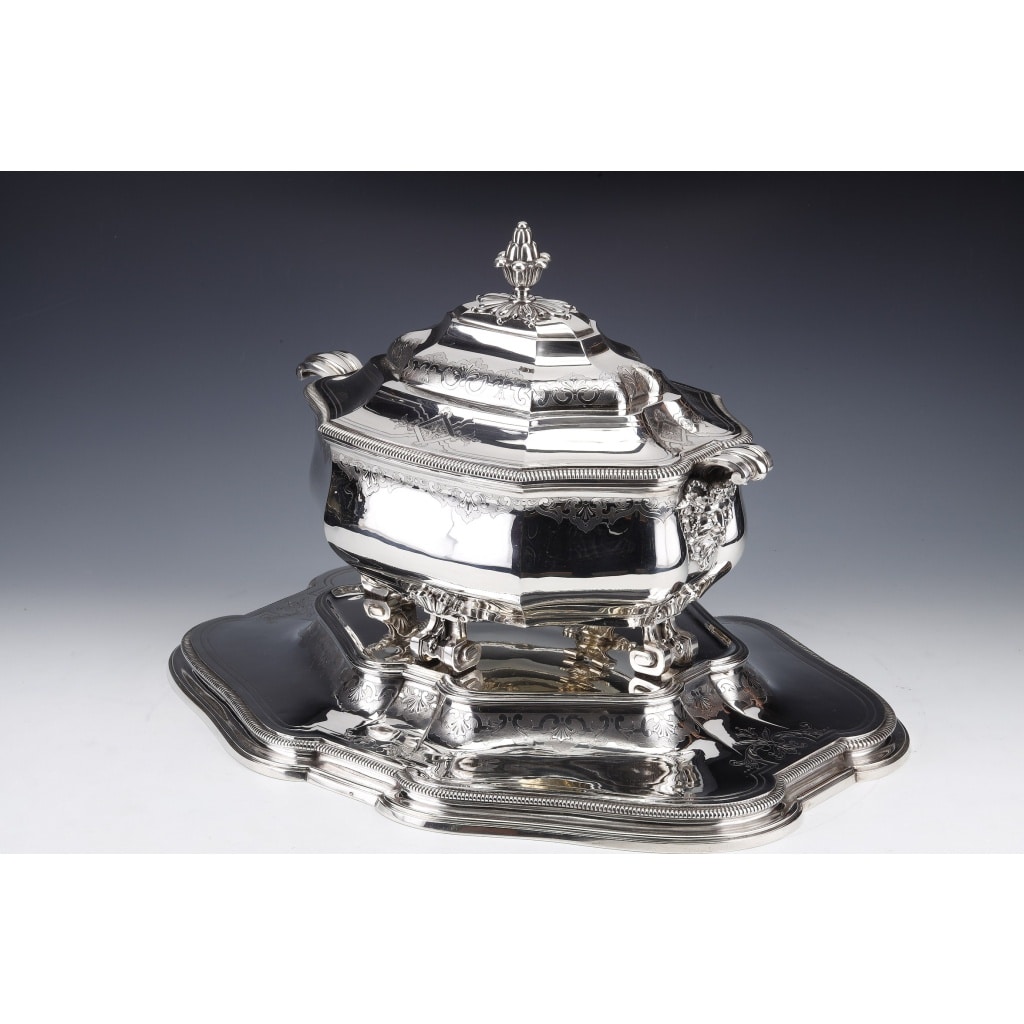 Important solid silver centerpiece represented by a covered soup tureen mounted on four scrolled feet and placed on its display stand. Rich decoration of Bérain engravings, edging of gadroons, the side handles in the form of volutes surmount mascarons, the double ogee lid has its handle in the form of a pine cone. The oval, scalloped display stand, bordered by a gadroon, has the same Bérain engraving decoration on the upright. Dimensions: Soup tureen length 39,5 cm - width 23 cm - height 28 cm Display stand: length 51 cm - width 40 cm - height 5,5 cm Total weight: 6700 grams approx. Material: 1st title silver 950/°°° Hallmark: MINERVE Period: FINISHED XIXth Goldsmith: A. AUCOC Origin: FRANCE/PARIS