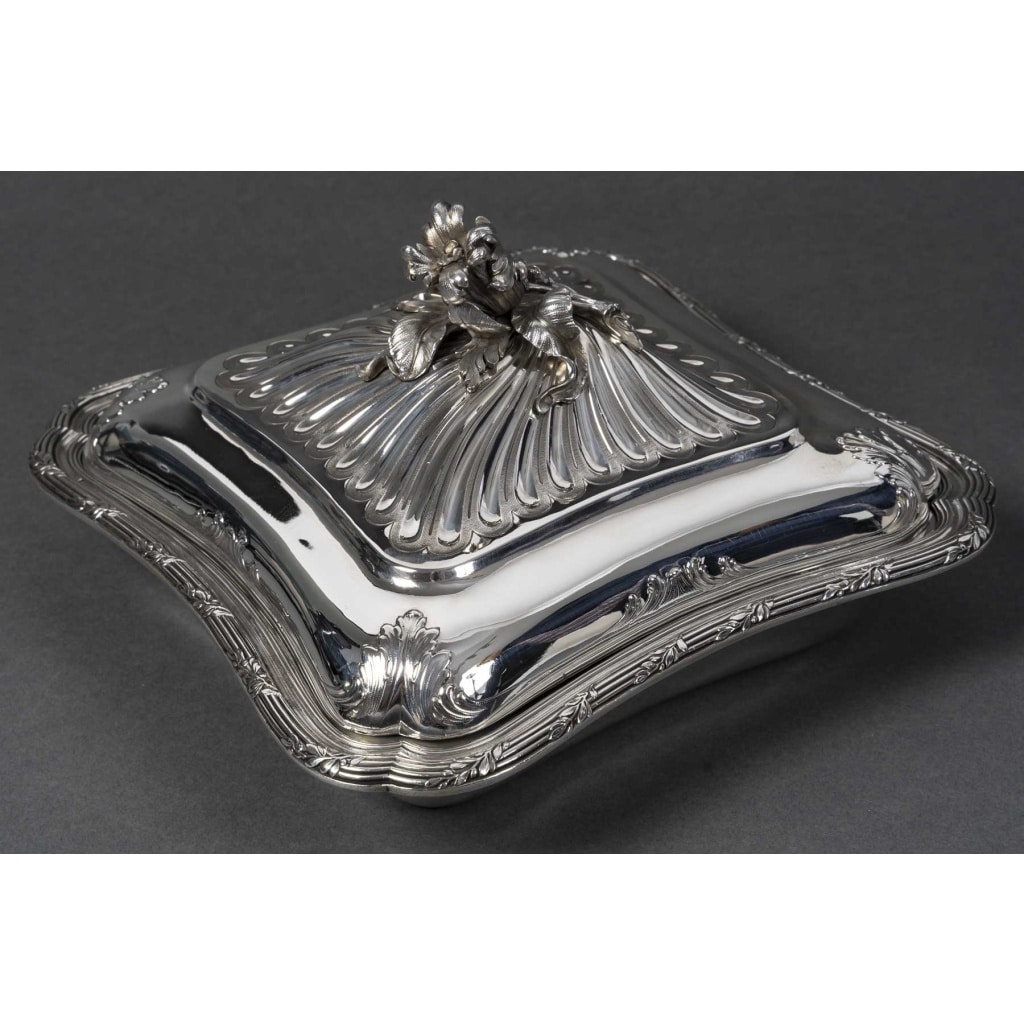 CARDEILHAC - LÉGUMIER COUVERT EN ARGENT MASSIF XIXe Légumier carré à angles adoucis de style Louis XV, orné d’une frise de coquilles et de guirlandes feuillagées finement ciselées. Le couvercle à côtes torsadées est sommé d’une prise en forme de bouquet de fleurs et feuillages naturalistes. Monogramme gravé sur la panse. Dimensions : 24 × 24 cm - Base : 17,5 × 17,5 cm - Hauteur : 13 cm Matière : Argent 1er titre 950/1°°° Poids brut : 1 490 g Poinçon : MINERVE Epoque : FIN XIXe Orfèvre : CARDEILHAC A PARIS Origine : FRANCE/PARIS Bibliographie : Maison Cardeilhac (1804-1951) Fondée en 1804 par Antoine-Vital Cardeilhac, la maison Cardeilhac s’est rapidement imposée parmi les orfèvres parisiens les plus prestigieux du XIXᵉ siècle. Réputée pour la qualité de ses ciselures et ses créations à la fois classiques et novatrices, elle excelle particulièrement dans la réinterprétation des styles Louis XV et Rocaille, prisés sous le Second Empire et la Troisième République. Sous la direction successive de ses descendants, Cardeilhac participe aux grandes Expositions Universelles de Paris (notamment 1867, 1878, 1889 et 1900) où elle obtient plusieurs médailles et distinctions. Elle développe des services de table raffinés, des pièces de prestige et des orfèvreries d’exception, souvent commandées par une clientèle aristocratique et bourgeoise. En 1951, la maison Cardeilhac est rachetée par Christofle, qui perpétue une partie de ses modèles et conserve l’héritage de son savoir-faire. Aujourd’hui, les pièces signées Cardeilhac sont très recherchées pour leur élégance et leur qualité de fabrication, illustrant l’âge d’or de l’orfèvrerie française.