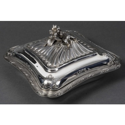 CARDEILHAC - LÉGUMIER COUVERT EN ARGENT MASSIF XIXe Légumier carré à angles adoucis de style Louis XV, orné d’une frise de coquilles et de guirlandes feuillagées finement ciselées. Le couvercle à côtes torsadées est sommé d’une prise en forme de bouquet de fleurs et feuillages naturalistes. Monogramme gravé sur la panse. Dimensions : 24 × 24 cm - Base : 17,5 × 17,5 cm - Hauteur : 13 cm Matière : Argent 1er titre 950/1°°° Poids brut : 1 490 g Poinçon : MINERVE Epoque : FIN XIXe Orfèvre : CARDEILHAC A PARIS Origine : FRANCE/PARIS Bibliographie : Maison Cardeilhac (1804-1951) Fondée en 1804 par Antoine-Vital Cardeilhac, la maison Cardeilhac s’est rapidement imposée parmi les orfèvres parisiens les plus prestigieux du XIXᵉ siècle. Réputée pour la qualité de ses ciselures et ses créations à la fois classiques et novatrices, elle excelle particulièrement dans la réinterprétation des styles Louis XV et Rocaille, prisés sous le Second Empire et la Troisième République. Sous la direction successive de ses descendants, Cardeilhac participe aux grandes Expositions Universelles de Paris (notamment 1867, 1878, 1889 et 1900) où elle obtient plusieurs médailles et distinctions. Elle développe des services de table raffinés, des pièces de prestige et des orfèvreries d’exception, souvent commandées par une clientèle aristocratique et bourgeoise. En 1951, la maison Cardeilhac est rachetée par Christofle, qui perpétue une partie de ses modèles et conserve l’héritage de son savoir-faire. Aujourd’hui, les pièces signées Cardeilhac sont très recherchées pour leur élégance et leur qualité de fabrication, illustrant l’âge d’or de l’orfèvrerie française.