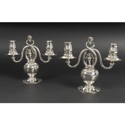 Georg JENSEN Paire de candélabres en argent massif, XXe ART DECO Superbe paire de candélabres à deux bras de lumière signée Georg Jensen, l’un des plus grands orfèvres du XXe siècle. Réalisés en argent massif (sterling silver), ces candélabres présentent un élégant piétement sphérique soutenant deux bras sinueux, ornés de binets évasés décorés d’un motif de baies en relief. La volute sommitale, typique du style naturaliste de Jensen, apporte équilibre et raffinement à l’ensemble. Dimensions : Hauteur 22 cm - largeur 25 cm Poids : 2 Kg 285 Epoque : Début XXè Orfèvre : Georg Jensen Poinçons : Georg Jensen – Denmark – Sterling - 324 État : excellent, argent légèrement poli, poinçons bien visibles. Ces pièces emblématiques du design scandinave début XXe siècle allient pureté des lignes, qualité artisanale et inspiration organique, caractéristiques des créations de Georg Jensen. Bibliographie : – Janet Drucker, Georg Jensen : A Tradition of Splendid Silver, New York, 1997, modèles similaires reproduits. Candélabre N°324 conçu par GEORG JENSEN 1919. Avec l'aimable autorisation de Georg Jensen/Royal Copenhagen. Un exemple similaire se trouve dans les collections du METROPOLITAIN MUSEUM OF ART NEW YORK Ces candélabres illustrent parfaitement l’esthétique organique du maître orfèvre Georg Jensen (1866-1935), inspirée par la nature et caractéristique du style Arts & Crafts scandinave.
