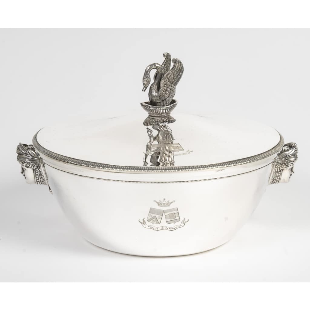 Orfèvre ANTOINE BOUILLET - Légumier couvert en argent massif Epoque 1er Empire Superbe légumier rond en argent massif, à panse lisse ornée de deux appliques en relief figurant des têtes féminines de style néo-classique, encadrées de guirlandes et de motifs floraux. Le couvercle, ceint d’une frise de filets torsadés et de perles finement ciselés, est surmonté d’une prise spectaculaire en forme de cygne aux ailes déployées, posé sur une coupe, emblème emblématique de l’esthétique impériale. La panse est gravée d’armoiries timbrées d’une couronne de marquis, accompagnées de la devise « Nullus Extinguetur ». Très bon état général, légères traces d’usage. Doublure postérieure en argent massif éxécutée par l'orfèvre COSSON CORBY Dimensions : Hauteur totale : env. 17,5 cm - Diamètre : env. 25 cm Poids total : 1 Kg840 Matière : Argent 1er titre 950/1°°° Poinçon de garantie au 1er coq (Paris, 1798–1809) Poinçon de maître orfèvre : ANTOINE BOUILLET Origine : France/PARIS L’orfèvre Antoine Bouillet Orfèvre parisien actif sous le Consulat et le Premier Empire, Bouillet s’inscrit dans la lignée des grands maîtres de l’orfèvrerie néo-classique. Ses pièces, destinées à une clientèle aristocratique et bourgeoise, se distinguent par leur sobriété architecturale et l’emploi de figures inspirées de l’Antiquité. Le cygne sous l’Empire Symbole de majesté, fidélité et grâce, le cygne devint sous Napoléon un motif récurrent des arts décoratifs. L’impératrice Joséphine en fit un emblème personnel, particulièrement visible à Malmaison, où il orne meubles, tissus et argenterie. Sur ce légumier, le cygne sculpté exprime toute la grandeur du style impérial, où l’animal acquiert une portée décorative et politique. Le légumier dans l’art de la table impérial Pièce centrale des services de table, le légumier couvert illustre la codification des banquets officiels : les légumes, servis en argent, étaient mis en valeur dans des pièces au décor monumental. Ornés d’armoiries et de symboles, ils affirmaient la puissance et le rang de leurs propriétaires.