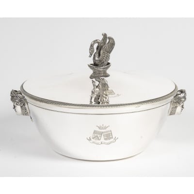 Orfèvre ANTOINE BOUILLET - Légumier couvert en argent massif Epoque 1er Empire Superbe légumier rond en argent massif, à panse lisse ornée de deux appliques en relief figurant des têtes féminines de style néo-classique, encadrées de guirlandes et de motifs floraux. Le couvercle, ceint d’une frise de filets torsadés et de perles finement ciselés, est surmonté d’une prise spectaculaire en forme de cygne aux ailes déployées, posé sur une coupe, emblème emblématique de l’esthétique impériale. La panse est gravée d’armoiries timbrées d’une couronne de marquis, accompagnées de la devise « Nullus Extinguetur ». Très bon état général, légères traces d’usage. Doublure postérieure en argent massif éxécutée par l'orfèvre COSSON CORBY Dimensions : Hauteur totale : env. 17,5 cm - Diamètre : env. 25 cm Poids total : 1 Kg840 Matière : Argent 1er titre 950/1°°° Poinçon de garantie au 1er coq (Paris, 1798–1809) Poinçon de maître orfèvre : ANTOINE BOUILLET Origine : France/PARIS L’orfèvre Antoine Bouillet Orfèvre parisien actif sous le Consulat et le Premier Empire, Bouillet s’inscrit dans la lignée des grands maîtres de l’orfèvrerie néo-classique. Ses pièces, destinées à une clientèle aristocratique et bourgeoise, se distinguent par leur sobriété architecturale et l’emploi de figures inspirées de l’Antiquité. Le cygne sous l’Empire Symbole de majesté, fidélité et grâce, le cygne devint sous Napoléon un motif récurrent des arts décoratifs. L’impératrice Joséphine en fit un emblème personnel, particulièrement visible à Malmaison, où il orne meubles, tissus et argenterie. Sur ce légumier, le cygne sculpté exprime toute la grandeur du style impérial, où l’animal acquiert une portée décorative et politique. Le légumier dans l’art de la table impérial Pièce centrale des services de table, le légumier couvert illustre la codification des banquets officiels : les légumes, servis en argent, étaient mis en valeur dans des pièces au décor monumental. Ornés d’armoiries et de symboles, ils affirmaient la puissance et le rang de leurs propriétaires.